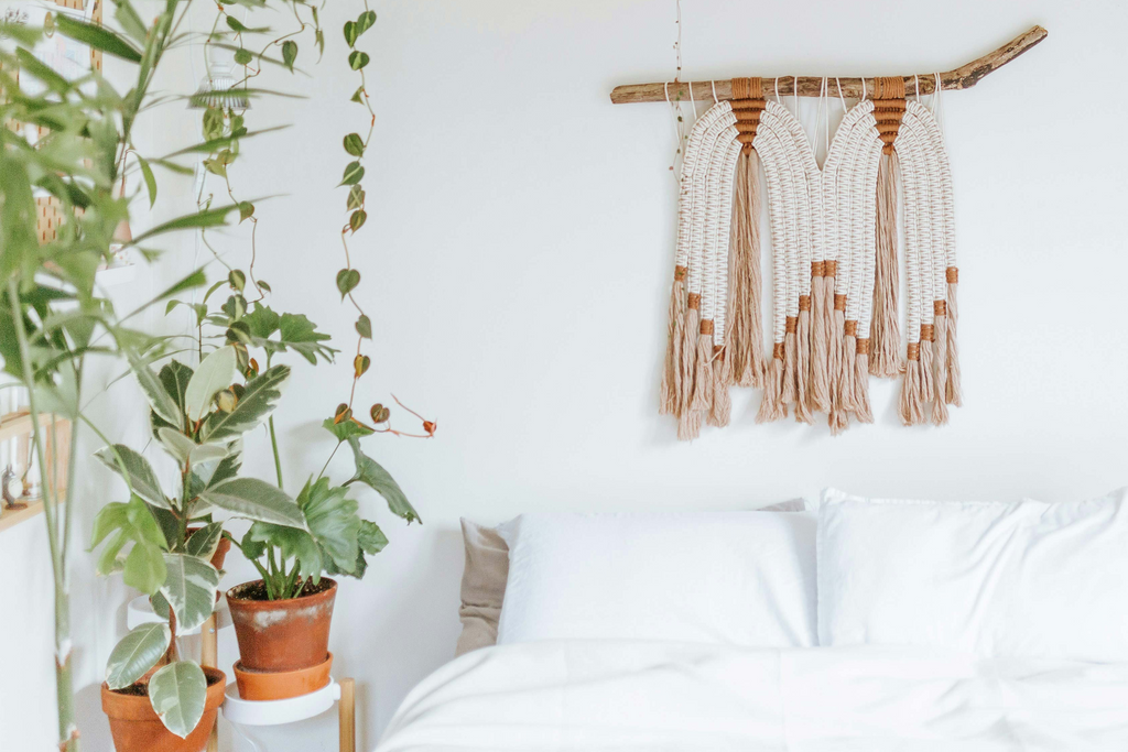 Foto de quarto em tons brancos, com plantas ao lado da cama e painel em macramé por cima da cama.